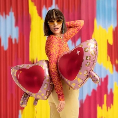 Mulher com camisola rosa de padrão cor-de-laranja e óculos de sol amarelos segurando balões de coração cor-de-rosa com padrão dourado e vermelho