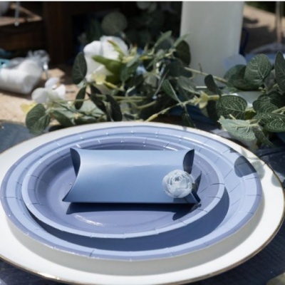 Prato branco com borda dourada, prato azul e embalagem de presente azul numa mesa decorada com folhas verdes