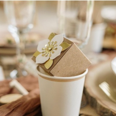 Copo de papel branco com caixa de presente decorada com flor branca e dourada numa mesa posta