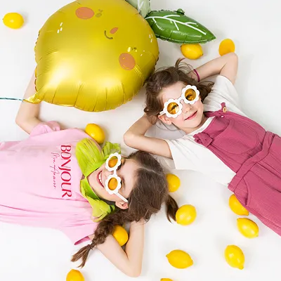 Duas meninas deitadas com roupas coloridas e óculos em formato de flor rodeadas por limas amarelas e balão amarelo em forma de lima.