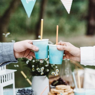 Duas mãos brindando com copos azuis num ambiente exterior decorado com mesa e árvores ao fundo