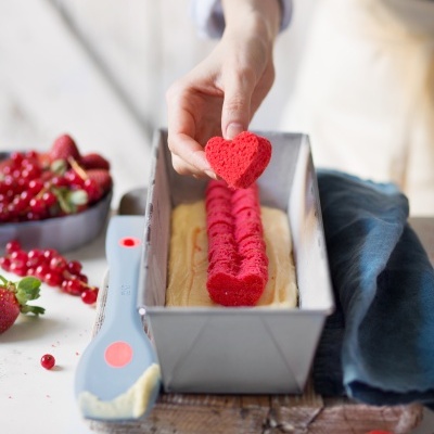 Bolachas vermelhas em forma de coração sobre massa em forma de metal retangular com frutos vermelhos e espátula ao lado