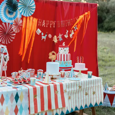 Mesa de festa de aniversário decorada com mesa com toalha colorida, bolo, chapéus e faixa HAPPY BIRTHDAY