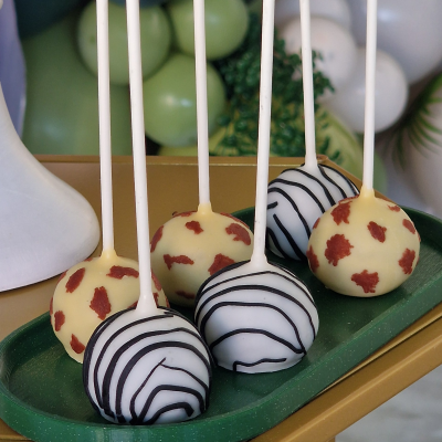 Cake pops decorados em bandeja verde sobre mesa dourada.