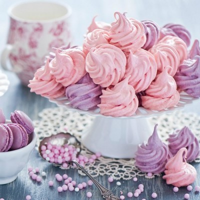 Suspiros rosa e lilás em prato branco com toalha rendada sobre mesa cinza.