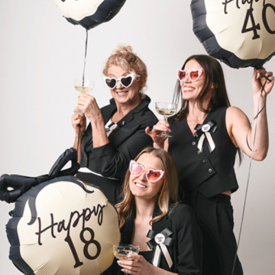 Três mulheres segurando balões redondos de festa com textos de aniversários e óculos de sol em forma de coração.