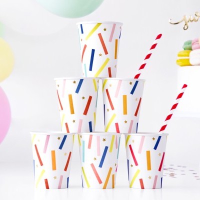 Seis copos de papel com riscas coloridas e dois canudos, junto a bolo branco com macarons e balões ao fundo