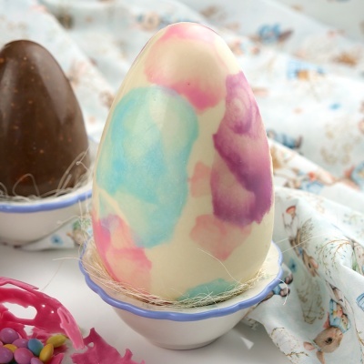 Ovo de chocolate branco com manchas coloridas numa tigela e ovo castanho ao fundo