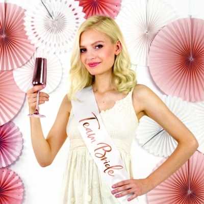 Mulher com vestido branco e faixa 'Team Bride' segurando taça de copo metálico rosa em fundo decorado com leques de papel