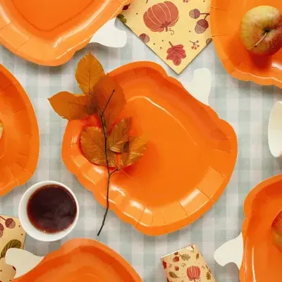 Pratos e copos de papel laranja com decoração de outono e maçãs sobre uma mesa xadrez cinza e branco