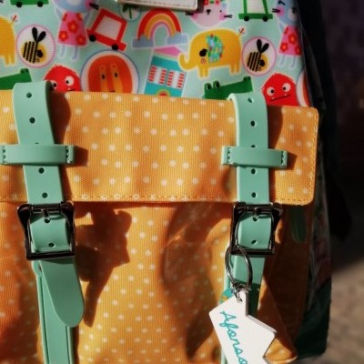 Mochila infantil colorida com bolso amarelo, alças verde-água e etiqueta branca com texto