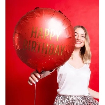 Balão vermelho metálico redondo com texto dourado HAPPY BIRTHDAY numa mão segurada por mulher com roupa branca e saia brilhante