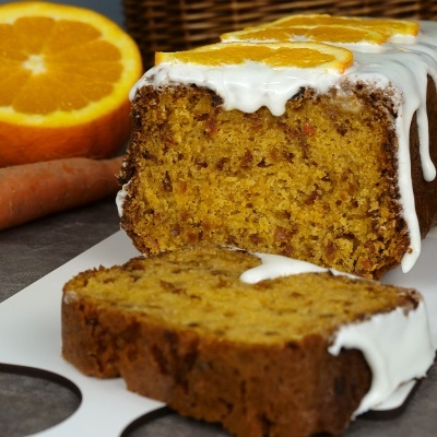 Bolo amarelo com glacê branco e fatias de laranja sobre uma superfície escura com cenoura e meia laranja ao fundo