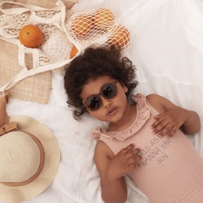 Criança com óculos de sol deitada em fundo branco com chapéu de palha e laranjas numa rede