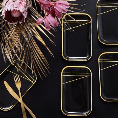 Pratos retangulares pretos com bordas douradas e talheres dourados sobre mesa preta com flores secas