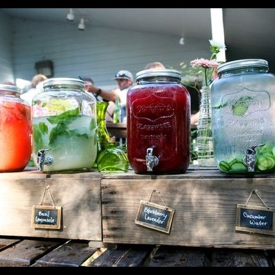 Quatro dispensadores de bebidas em vidro com torneiras, sobre um suporte de madeira, com etiquetas de quadro-negro e ambiente exterior com pessoas ao fundo