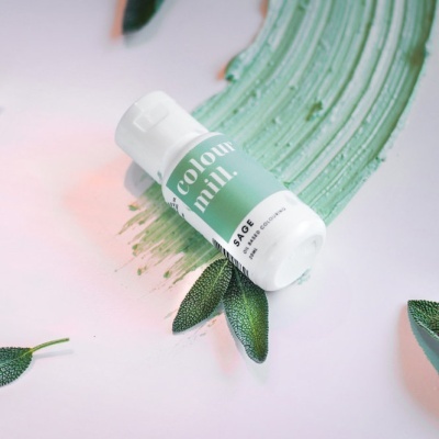 Frasco branco de produto com rótulo verde 'colour mill.' e 'SAGE' com folhas verdes e mancha verde ao fundo
