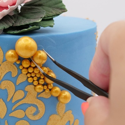 Bolo azul com decoração dourada e esferas douradas sendo ajustadas por pinças pretas