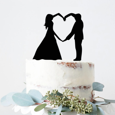 Topo de bolo de casamento com silhueta de noiva e noivo em preto, bolo branco e folhas verdes decorativas.