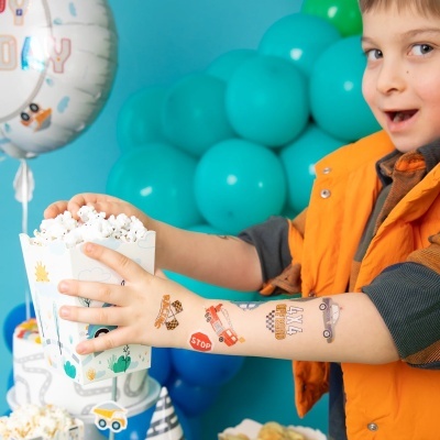 Menino com colete laranja segurando caixa branca de pipocas com balões azuis ao fundo