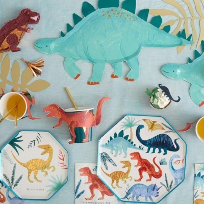 Conjunto de utensílios de mesa com tema de dinossauros para crianças em cores variadas sobre mesa azul clara com folhas. 