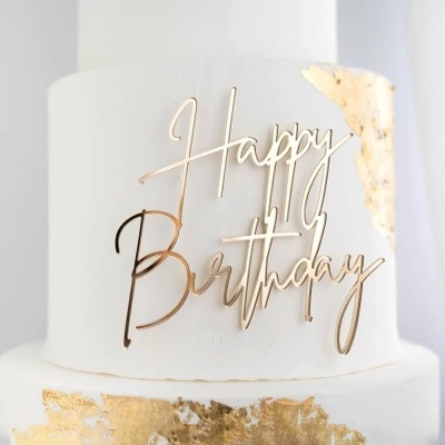 Bolo branco de três andares com topper Happy Birthday dourado e folhas douradas decorativas.