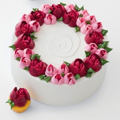 Bolo branco decorado com flores de glacê vermelhas e rosas, com um cupcake ao lado.