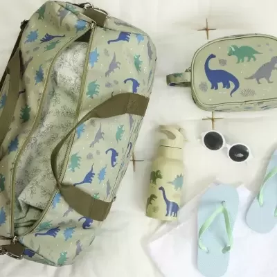 Conjunto de acessórios com padrão de dinossauros em tons de verde e azul, incluindo mala, garrafa, nécessaire, chinelos e óculos de sol