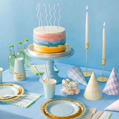 Mesa de festa com bolo multicolorido, copos, pratos, chapéus de festa e balões em papel