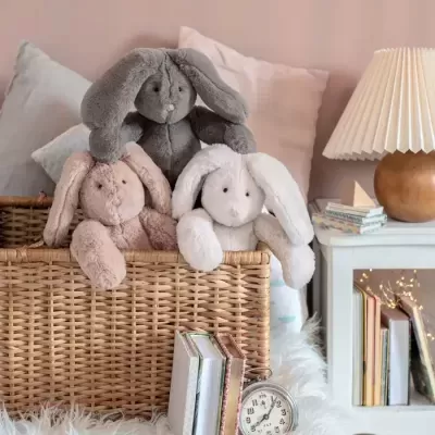 Três coelhos de peluche numa cesta de verga com almofadas coloridas e mesa branca com candeeiro