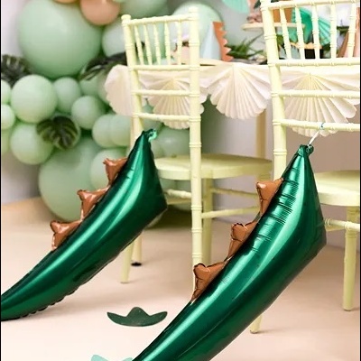 Balões metálicos verdes em forma de patas de dinossauro amarrados a cadeiras claras numa festa decorada com balões pastel e letras 'HAPPY'.