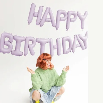 Balões lilases com as palavras HAPPY BIRTHDAY e pessoa agachada com camisola verde e botas amarelas