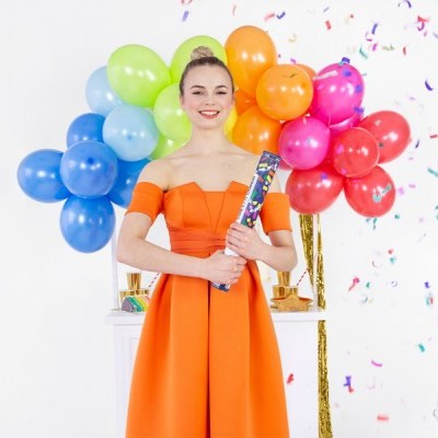 Mulher jovem vestindo vestido laranja segura tubo de confetes com balões coloridos e confetes ao fundo