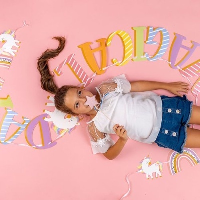 Menina deitada com blusa branca e saia azul sobre fundo rosa com decoração de unicórnios e letras coloridas