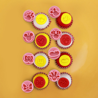 Dez bolos pequenos em amarelo, vermelho e branco com carimbos e cortadores rosa com formas de personagens animados sobre fundo amarelo.
