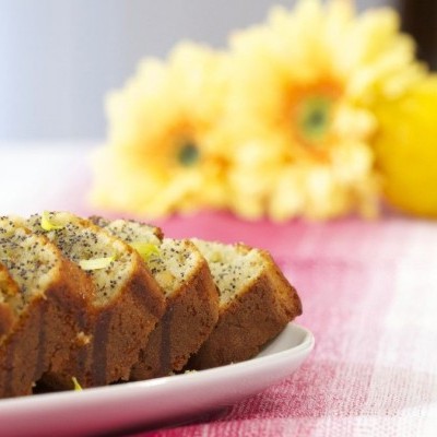 Bolo fatiado com sementes de papoila e limão num prato branco com limões e flores amarelas ao fundo