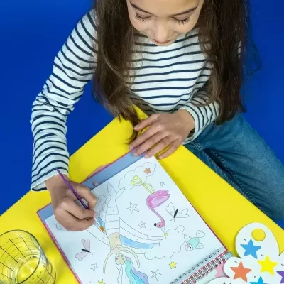 Caderno de colorir aberto sobre mesa amarela com criança a pintar desenhos com pincel