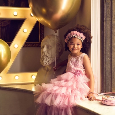 Menina com vestido rosa e coroa de flores segurando balão dourado junto a busto e letra Z luminosa