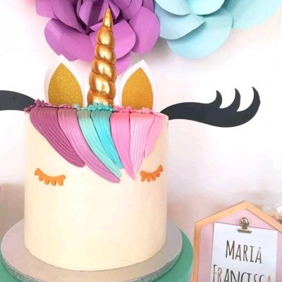 Bolo unicórnio com chifre dourado e decoração colorida, placa com nome 'MARÍA FRANCISCA' ao lado e flores de papel no fundo