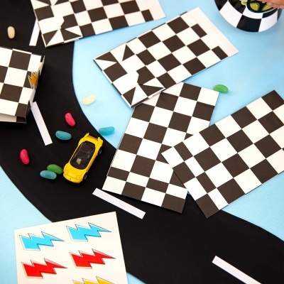 Envelopes com padrão xadrez, carrinho de brinquedo amarelo, estrada de papel preta, balas coloridas e autocolantes coloridos sobre fundo azul.