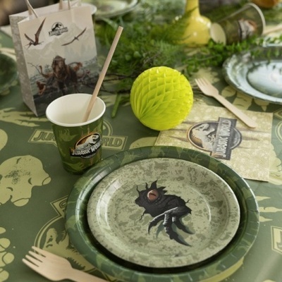 Conjunto de artigos descartáveis Jurassic World com pratos, copo, guardanapo e talheres sobre toalha verde com tema de dinossauros