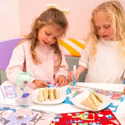 Duas crianças a desenhar e comer sanduíches em mesa com garrafa de água colorida