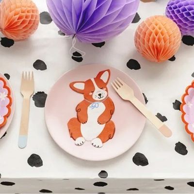 Prato rosa com desenho de cão, talheres de madeira e decoração em papel laranja e roxo sobre toalha branca com manchas pretas