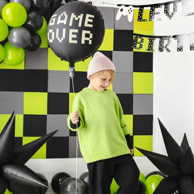 Criança com balão de festa 'GAME OVER' num ambiente decorado com balões coloridos e painel quadriculado.