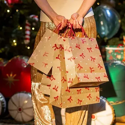 Pessoa com sacos de papel castanho com laços vermelhos e calças douradas junto a decoração de Natal