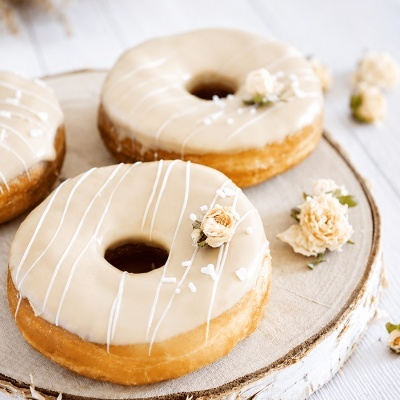 Três donuts com cobertura branca e decorados com pequenas flores creme sobre uma tábua de madeira clara.