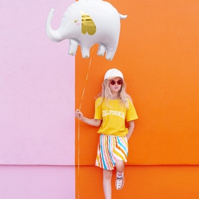 Criança com roupa colorida e balão elefante junto a parede laranja e rosa