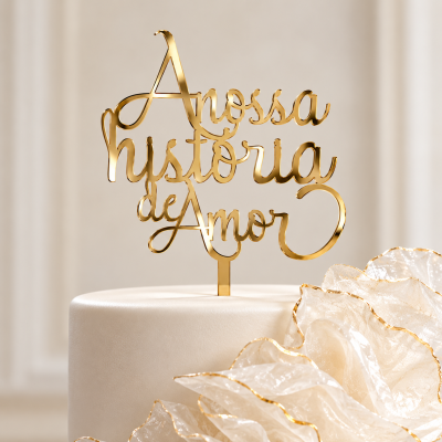 Topo de bolo dourado com texto A nossa história de Amor sobre bolo branco com decoração de pétalas douradas