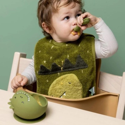 Bebé com babete de dinossauro a usar colher e tigela verdes em cadeira de alimentação