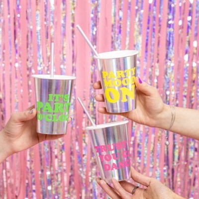 Três copos metálicos com textos coloridos e canudos, frente a um fundo de fitas brilhantes cor-de-rosa e lilás